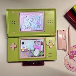 May include: Lime green Nintendo DS handheld game console with two screens showing colourful anime-style graphics. Includes a pink stylus, game cartridge, and a pink battery cover. The top screen reads "Kanroji Mitsuri".
