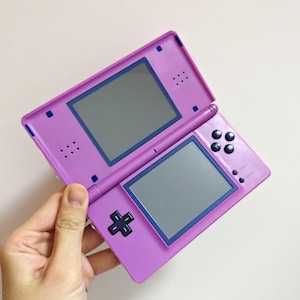 May include: A purple handheld gaming console with two screens and a stylus. The console features a directional pad, action buttons, and a power button. The screens are framed in blue. The console is open, revealing the screens.