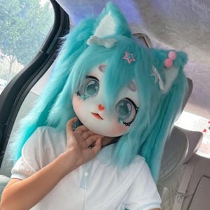 Può includere: Una maschera da gatto in stile anime blu chiaro con grandi occhi espressivi e lunghi capelli. La maschera presenta un piccolo cuore rosso sul naso ed è adornata da una stella e pompon rosa. La persona indossa una polo bianca.