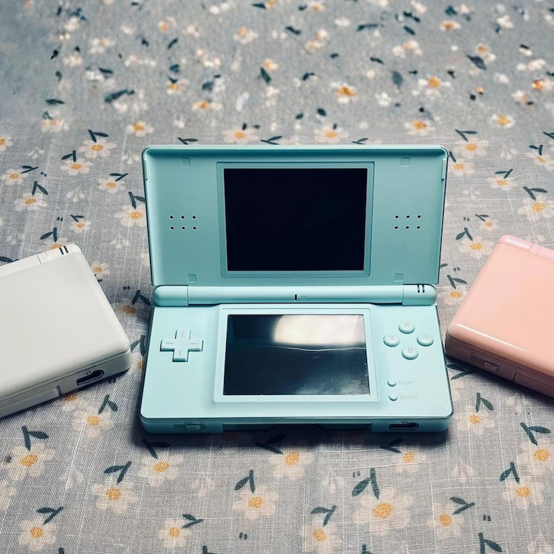 Puede incluir: Una consola de juegos port&aacute;til Nintendo DS azul claro est&aacute; abierta, mostrando dos pantallas. Una Nintendo DS blanca y otra rosa est&aacute;n a cada lado. Las consolas est&aacute;n sobre una superficie con estampado floral.