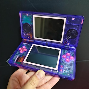 May include: A handheld video game console with a translucent purple plastic shell. The device features two screens, a directional pad, and four action buttons in bright pink. Internal circuit boards and wiring are visible through the casing.