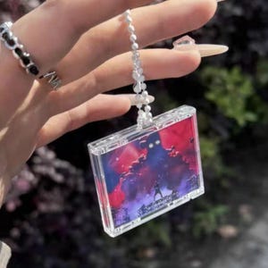 Può includere: Portachiavi rettangolare trasparente con una scena di concerto all'interno. L'immagine presenta una persona sul palco con uno sfondo rosso, viola e blu. Il portachiavi è attaccato a una catena di perline argentate.