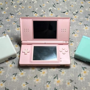 Puede incluir: Una consola de juegos port&aacute;til Nintendo DS rosa est&aacute; abierta, mostrando dos pantallas negras. Tambi&eacute;n se ven una Nintendo DS blanca y una turquesa. La consola tiene una cruceta, botones y una ranura para l&aacute;piz. El fondo es una tela con estampado floral.