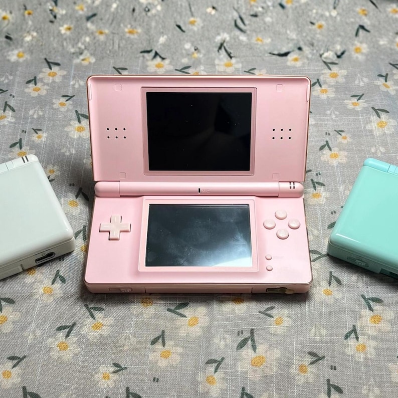 Puede incluir: Una consola de juegos port&aacute;til Nintendo DS rosa est&aacute; abierta, mostrando dos pantallas negras. Tambi&eacute;n se ven una Nintendo DS blanca y una turquesa. La consola tiene una cruceta, botones y una ranura para l&aacute;piz. El fondo es una tela con estampado floral.