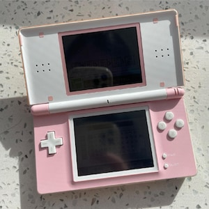 May include: A pink Nintendo DS handheld game console, open to display two screens. The top screen is white and pink, and the bottom screen is pink. The console has a directional pad, buttons, and a stylus.