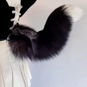 以下が含まれることがあります： ふわふわした質感の黒と白のフェイクファーの尻尾。 尻尾は主に黒で、白い先端とベースにシルバーグレーの毛皮のアクセントが付いています。 尻尾は黒い衣服に取り付けられています。