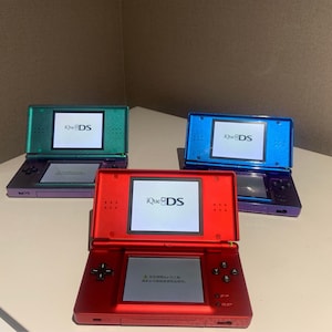 May include: Three iQue DS handheld gaming consoles in red, blue, and green. Each console is open, displaying a screen with the iQue DS logo. The consoles have a dual-screen design with a bottom screen and a top screen.