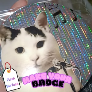 Op de afbeelding: Een ronde, zilveren badge met een holografisch oppervlak. Het toont een witte kat met zwarte vlekken die een microfoon vasthoudt. De tekst "MAKE YOUR BADGE" is in roze en zwart. Muzieknoten zijn zichtbaar.