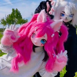 Può includere: Una persona indossa una testa di fursuit a tema gatto rosa con lunghi capelli rosa fluenti e zampe rosa abbinate. Il fursuit ha grandi occhi blu e un viso dettagliato. Un'altra persona indossa una testa di fursuit a tema gatto bianco con capelli bianchi.