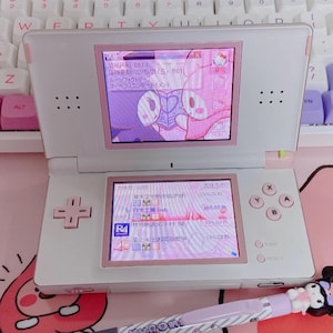 May include: A white Nintendo DS Lite handheld game console with pink trim. The screens show a pink interface with Japanese text and cartoon characters. A matching pen with a cartoon character is in the foreground.