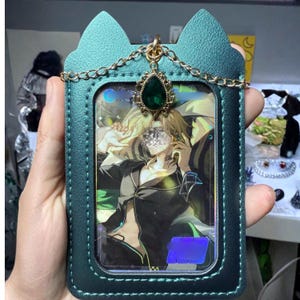 Op de afbeelding: Teal groene kaarthouder met kattenoordesign, gouden ketting en een decoratieve groene edelsteen. De houder bevat een kleurrijke afbeelding van een persoon. De houder wordt in een hand gehouden.