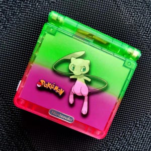 Puede incluir: Una Game Boy Advance SP verde y rosa con un gráfico de Mew Pokémon. La consola portátil tiene una parte superior verde y una cubierta de pantalla rosa y verde. El logotipo de Pokémon es visible, junto con el logotipo de Nintendo.