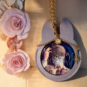 Op de afbeelding: Een lichtpaars, glinsterend konijnvormig accessoire met een gouden ketting. Het accessoire bevat een cirkelvormige afbeelding met een persoon en tekst. Een gouden sterbedel en een kleine bloembedel hangen aan de ketting. Roze papieren rozen staan op de achtergrond.