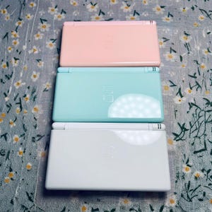 Puede incluir: Tres consolas de juegos port&aacute;tiles de colores pastel: rosa, turquesa y blanco. Cada consola est&aacute; cerrada, mostrando la mitad superior con la pantalla y los botones. Las consolas est&aacute;n apiladas, con la rosa en la parte superior.