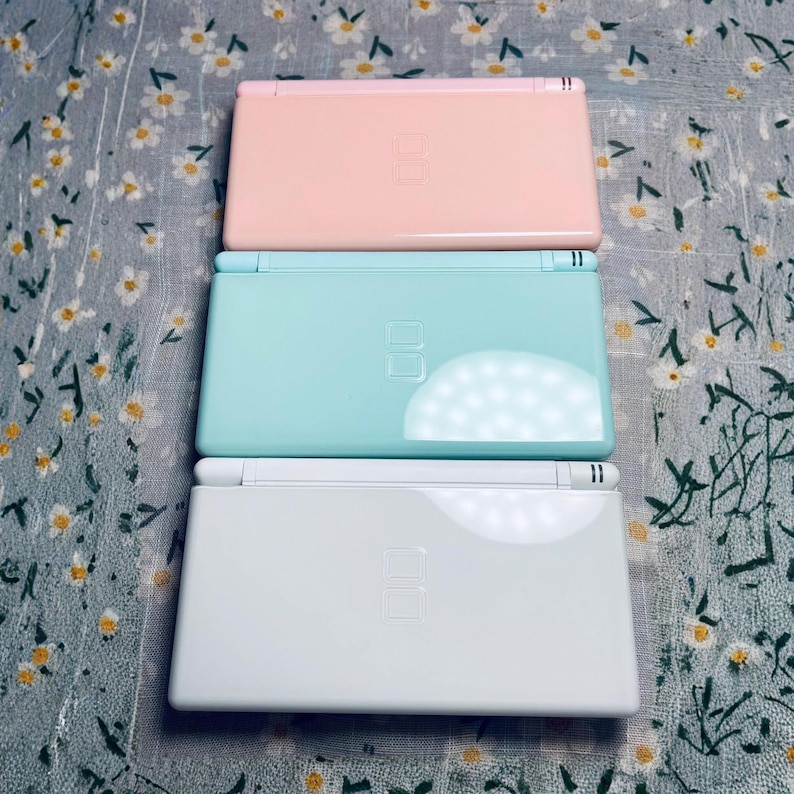 Puede incluir: Tres consolas de juegos port&aacute;tiles de colores pastel: rosa, turquesa y blanco. Cada consola est&aacute; cerrada, mostrando la mitad superior con la pantalla y los botones. Las consolas est&aacute;n apiladas, con la rosa en la parte superior.