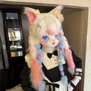 Può includere: Una maschera a forma di bambola con pelliccia bianca, accenti rosa e blu e grandi occhi blu. La maschera ha orecchie e capelli attaccati acconciati in trecce con nastri pastello. La bambola indossa un abito da cameriera bianco e nero con un papillon.