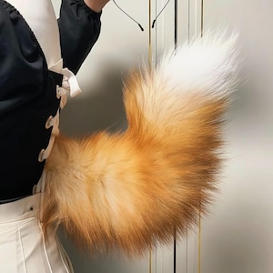 Può includere: Una soffice coda di volpe arancione e bianca. La coda è di un arancione vibrante alla base, che sfuma in un bianco morbido sulla punta. La coda è piena e sembra morbida. È un accessorio di costume.