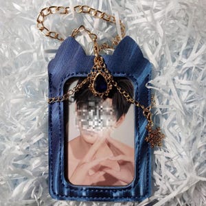 Op de afbeelding: Een marineblauwe ID-kaarthouder met een gouden ketting en een decoratieve sneeuwvlokbedel. De houder heeft een kattenoordesign aan de bovenkant en een gouden ketting met een blauwe edelsteen. De houder bevat een foto.