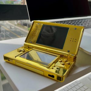 May include: A gold-colored Nintendo DS handheld game console. The console is open, revealing two screens. The top screen is black, and the bottom screen is reflective. The buttons and exterior are all gold.