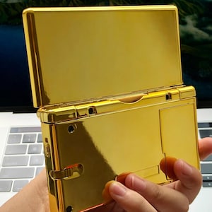 May include: A gold-colored handheld gaming console with a reflective, glossy finish. The top screen is open, revealing the interior. The device is held in a person's hand, showcasing its size and design. The console has a sleek, modern aesthetic.