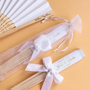 Peut inclure: Un éventail blanc pliable avec une structure en bambou et l'inscription Better Together, accompagné de deux éventails pliés dans des pochettes en organza. Chaque éventail présente un nœud en satin blanc, une perle et un pompon rose pâle avec le texte Jack & Linda 10.15.2027.