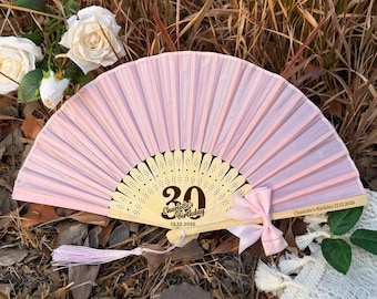 May include: A light pink folding hand fan with a wooden frame featuring laser-cut details. The fan displays the text 30, Charlotte's Birthday, and 12.12.2026 in dark brown ink. A small pink ribbon bow and a matching tassel are attached to the frame.