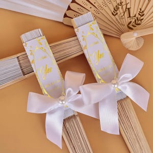 Peut inclure: Deux éventails pliés avec des baleines en bois clair et un tissu blanc. Chaque éventail comporte un fourreau en papier blanc avec le texte doré "He Said Yes", "Jack & Linda" et une date. Un nœud en satin blanc avec une perle est fixé.