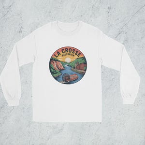 May include: White long-sleeve shirt featuring a circular graphic of La Crosse, Wisconsin. The design includes a river scene with a boat, cliffs, and the text "LA CROSSE WISCONSIN" in a circular arrangement. The shirt has a crew neck.