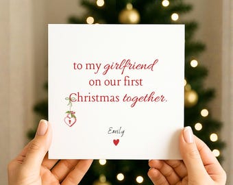 May include: A white card with red text that reads "to my girlfriend on our first Christmas together." The card has a small illustration of a heart-shaped lock and the name "Emily" with a red heart below. The card is held in front of a Christmas tree.