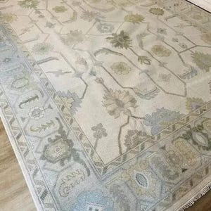 May include: A large, rectangular area rug with an intricate floral pattern in muted tones of cream, sage green, and pale blue. The rug features a detailed border and a central design of stylized flowers and vines, creating a vintage aesthetic.