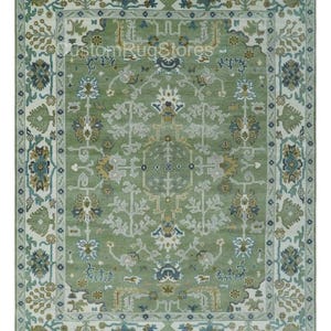 May include: A rectangular area rug with an intricate floral and geometric pattern. The rug features a central field of green with a detailed design in shades of blue, beige, and gray. A cream-colored border frames the design.