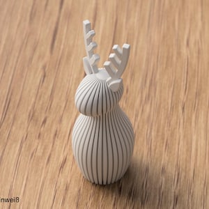 Pu&ograve; includere: Una statuetta di cervo tridimensionale in bianco opaco, costruita con lamelle verticali parallele. L'oggetto presenta un corpo arrotondato, una testa liscia e corna stilizzate con un design dentellato. Il testo "ELFENBEINWEISS" &egrave; stampato nell'angolo.