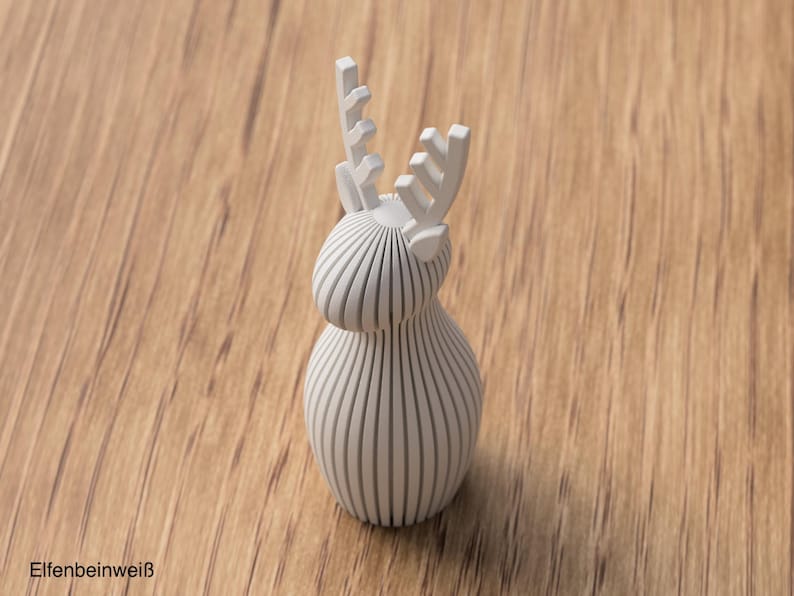 Pu&ograve; includere: Una statuetta di cervo tridimensionale in bianco opaco, costruita con lamelle verticali parallele. L'oggetto presenta un corpo arrotondato, una testa liscia e corna stilizzate con un design dentellato. Il testo "ELFENBEINWEISS" &egrave; stampato nell'angolo.