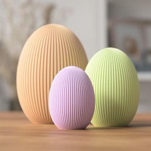 Könnte beinhalten: Drei dekorative, eiförmige Objekte mit vertikal geriffelten Texturen. Die Eier sind in den Farbtönen Pfirsich, Lavendel und Hellgrün gehalten. Sie sind auf einer Holzoberfläche angeordnet, mit einem unscharfen Hintergrund.
