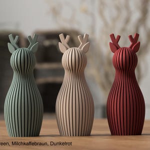 Pu&ograve; includere: Tre statuette di cervo in piedi con dettagli a lamelle verticali. La figura a sinistra &egrave; verde tenue, quella centrale &egrave; beige e quella a destra &egrave; rosso scuro. Ogni cervo ha un corpo arrotondato e corna stilizzate. Il testo Muted Green, Milchkaffeebraun, Dunkelrot &egrave; visibile in basso.