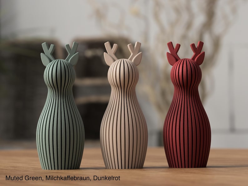 Pu&ograve; includere: Tre statuette di cervo in piedi con dettagli a lamelle verticali. La figura a sinistra &egrave; verde tenue, quella centrale &egrave; beige e quella a destra &egrave; rosso scuro. Ogni cervo ha un corpo arrotondato e corna stilizzate. Il testo Muted Green, Milchkaffeebraun, Dunkelrot &egrave; visibile in basso.