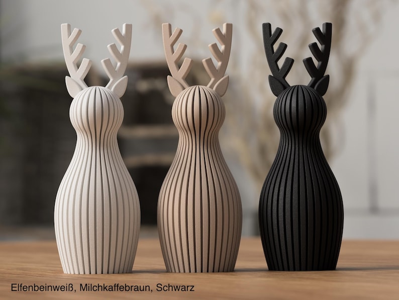 Pu&ograve; includere: Tre statuette di cervo in piedi con corpi a coste verticali e corna ramificate. Le figure sono di colore bianco avorio, marrone caff&egrave; latte e nero. Il testo in basso recita: ELFENBEINWEISS, MILCHKAFFEEBRAUN, SCHWARZ.