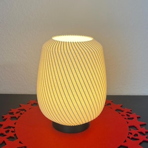 Könnte beinhalten: Zylindrische Tischlampe mit cremefarbenem, geripptem Schirm und schwarzem Sockel. Die Lampe steht auf einer roten, kreisförmigen Tischunterlage mit Sternaussparungen. Die Lampe ist beleuchtet und strahlt ein warmes Licht aus. Höhe ca. 25 cm.