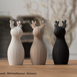Pu&ograve; includere: Tre statuette di cervo in piedi con una texture a coste verticali. I cervi sono color bianco avorio, beige chiaro e nero. Ogni statuetta presenta un corpo arrotondato e corna stilizzate. Il testo in basso recita Elfenbeinwei&szlig;, Milchkaffeebraun, Schwarz.