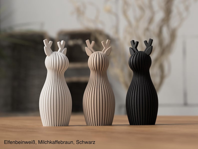 Pu&ograve; includere: Tre statuette di cervo in piedi con una texture a coste verticali. I cervi sono color bianco avorio, beige chiaro e nero. Ogni statuetta presenta un corpo arrotondato e corna stilizzate. Il testo in basso recita Elfenbeinwei&szlig;, Milchkaffeebraun, Schwarz.
