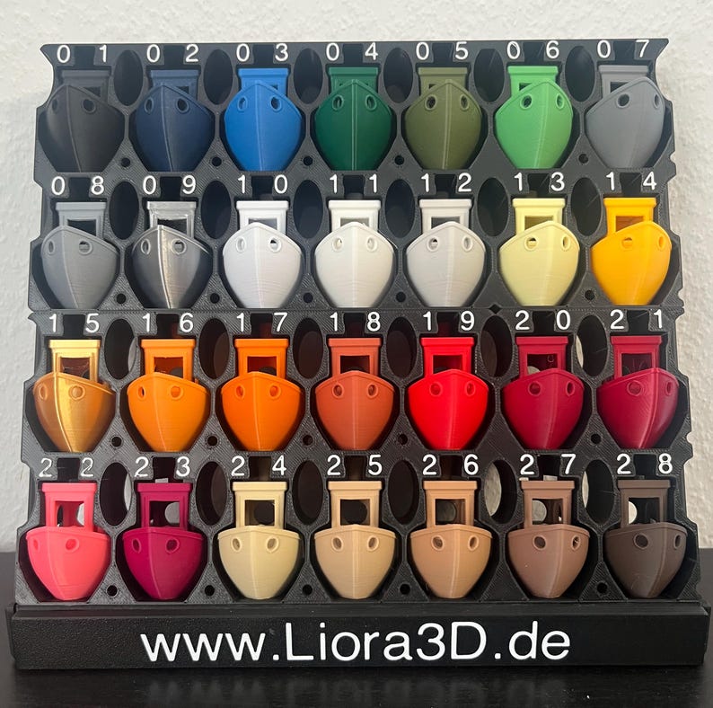 Pu&ograve; includere: Un espositore nero contenente ventinove piccoli modelli di rimorchiatori in plastica stampati in 3D in vari colori, numerati da 01 a 28. La base dell'espositore presenta il testo bianco www.Liora3D.de, mostrando un ampio spettro di tonalit&agrave;.