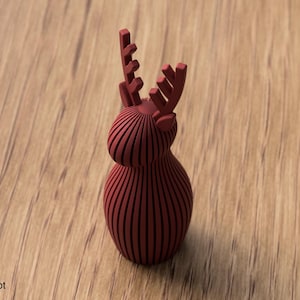 Pu&ograve; includere: Una statuetta di renna color rosso scuro con finitura opaca, costruita con una serie di lamelle verticali parallele. L'oggetto presenta un corpo e una testa arrotondati e bulbosi con due palchi stilizzati e ramificati verso l'alto.