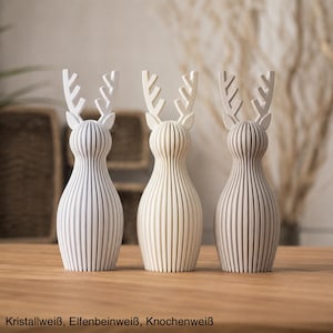 Pu&ograve; includere: Tre statuette di renne in piedi con texture a coste verticali. La figura a sinistra &egrave; bianco cristallo, quella centrale &egrave; avorio e quella a destra &egrave; bianco osso. Ognuna presenta palchi stilizzati e un corpo arrotondato. Testo: Kristallwei&szlig;, Elfenbeinwei&szlig;, Knochenwei&szlig;.