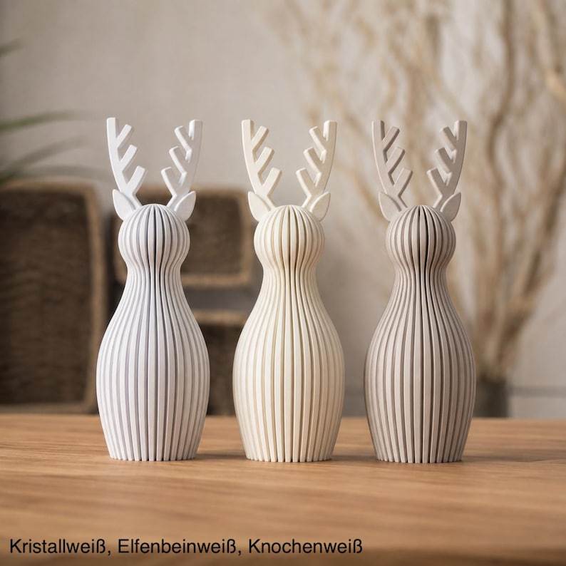 Pu&ograve; includere: Tre statuette di renne in piedi con texture a coste verticali. La figura a sinistra &egrave; bianco cristallo, quella centrale &egrave; avorio e quella a destra &egrave; bianco osso. Ognuna presenta palchi stilizzati e un corpo arrotondato. Testo: Kristallwei&szlig;, Elfenbeinwei&szlig;, Knochenwei&szlig;.
