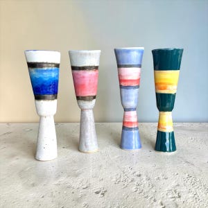 May include: Four ceramic vases in a distinctive, elongated form. Each vase showcases a different colour palette, including blue, pink, and green, with horizontal bands of colour. The vases are displayed on a light-coloured surface.