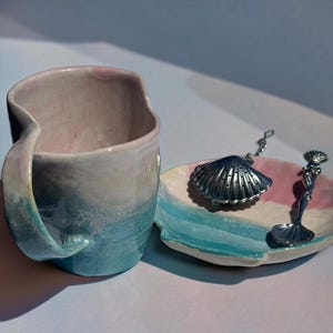 May include: A handmade ceramic mug and matching small plate featuring a gradient glaze of soft pink, cream, and turquoise blue. A silver-toned metal tea infuser shaped like a seashell and a matching spoon with a mermaid handle rest on the plate.