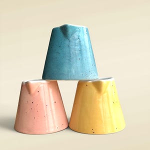 May include: Three ceramic jugs stacked in a pyramid. The jugs are in blue, pink, and yellow. Each has a spout and a speckled finish. The background is a plain, neutral colour.