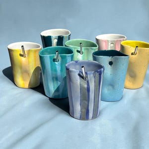 May include: A collection of eight ceramic tumblers in various colours including yellow, teal, light green, pink, blue, and purple. Each tumbler features an irregular, hand-formed shape, a smooth glazed finish, and a small hole near the rim holding a metal straw.