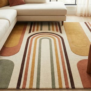 May include: A cream-colored area rug with a geometric design featuring arches and stripes in shades of brown, orange, yellow, and green. A beige sectional sofa and a rust-colored armchair are placed on the rug.