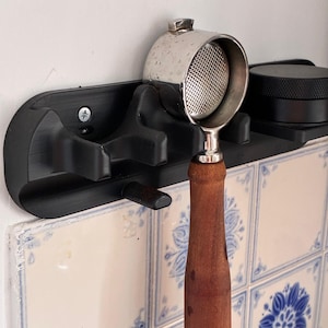 Peut inclure: Support mural noir pour porte-filtre de machine à expresso. Il maintient un porte-filtre argenté avec un manche en bois. Le support est fixé sur un mur blanc avec des carreaux à motifs floraux bleus.